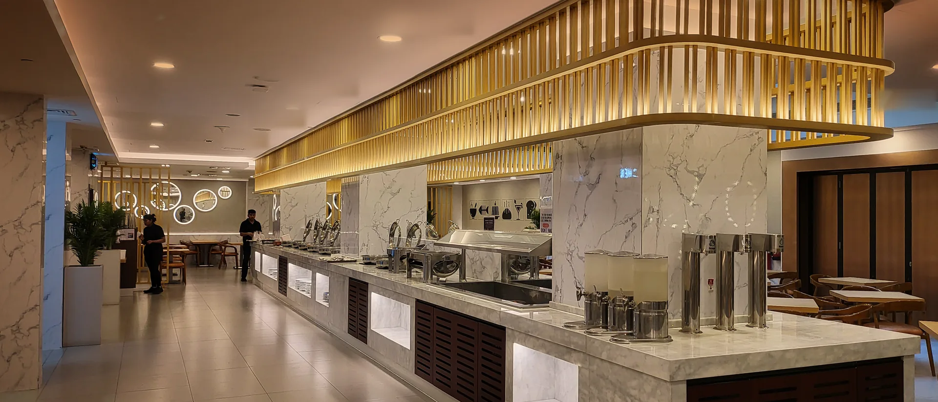 A food court counter under a warm gold slatted canopy, with marble counters and chafing dishes